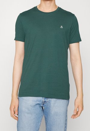 Mann trägt ein dunkelgrünes kurzärmeliges T-Shirt mit kleinem weißem "M O P" Logo auf der Brust, kombiniert mit hellblauen Jeans.