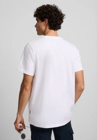 Camiseta de algodón blanca con mangas cortas, cuello redondo y corte relajado. Costuras visibles y un pequeño detalle de logo en el dobladillo.