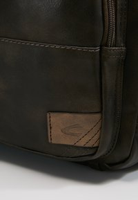 Braune Lederhandtasche mit strukturierter Oberfläche, ausgestattet mit einem Reißverschluss und einem gestickten Logopatch an der Seite. Glatte Oberfläche mit dezentem Glanz.