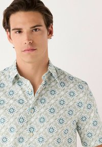 Camisa de botones de manga corta con un patrón floral verde y blanco con acentos azules, hecha de tela ligera. Diseño con cuello.