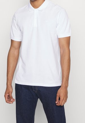 Mężczyzna w białej, gładkiej koszulce polo z krótkim rękawem i ciemnoniebieskich dżinsach na jednolitym tle.