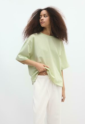 Femme aux cheveux bouclés et volumineux portant un t-shirt oversize vert clair et un pantalon blanc, soulevant légèrement le t-shirt avec la main.