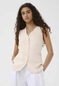Beige linneväst med V-ringning, fem framknappar och sidofickor. Texturerad tyg med avslappnad passform. Buren över vita byxor.