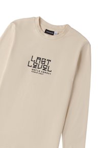 T-shirt beige a maniche lunghe con collo rotondo. La parte anteriore presenta la scritta 'LAST LEVEL' e 'WORLD FAMOUS SKATE BOUR' in testo grafico nero. Materiale in morbido cotone.