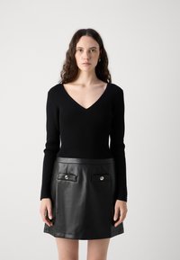 Pull en maille côtelée noir avec un col en V, associé à une mini-jupe en cuir noir dotée de deux poches à rabat et de boutons en argent.