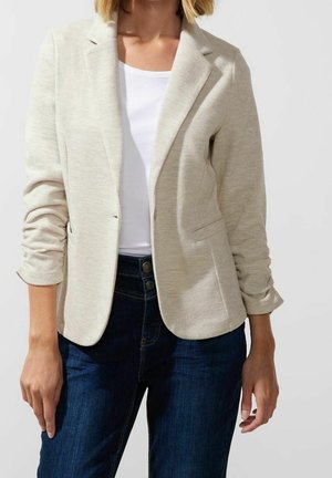 Kobieta w beżowej marynarce na białej koszuli i ciemnoniebieskich dżinsach na tle jasnego, jednolitego tła.