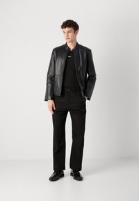 Black leather jacket with a high collar, worn over a black shirt and black trousers. Black shoes complete the outfit, emphasizing a monochromatic look.