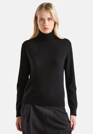 Femme blonde portant un pull noir à col roulé et un pantalon ample foncé, se tenant contre un fond blanc uni.