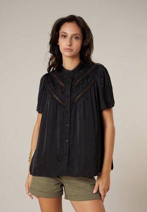 Femme portant une blouse noire ample à boutons avec des détails en dentelle et un short olive aux jambes retroussées, debout devant un fond neutre.