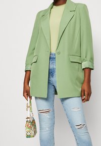 Blazer verde con rever a lancia e maniche arrotolate, abbinato a una maglietta verde chiaro e jeans blu strappati. Porta una borsa con motivo a disegni.