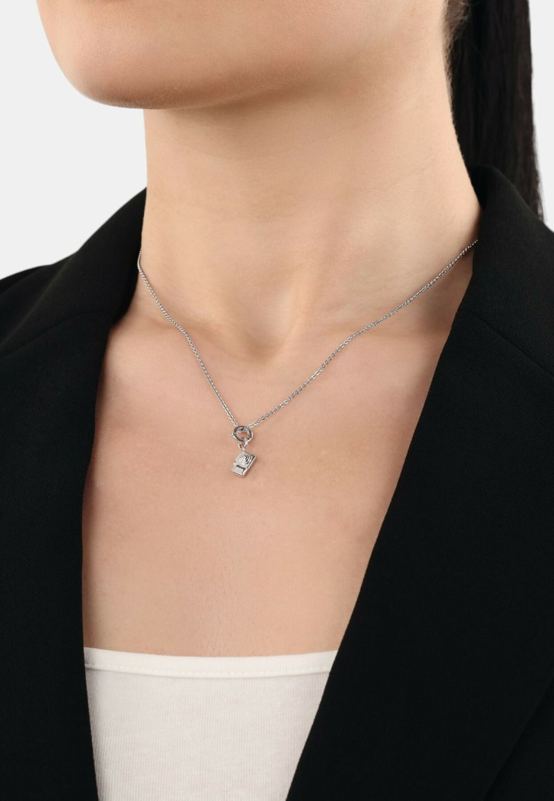 Collana d'argento con un piccolo ciondolo a forma di libro, dotata di una delicata catena e un accento a forma di rosa sul ciondolo. Indossata sopra una maglietta bianca.