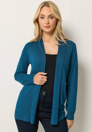 Blondynka w turkusowym, otwartym kardiganie na czarnej bluzce i ciemnych dżinsach, stojąca na jednolitym jasnym tle.