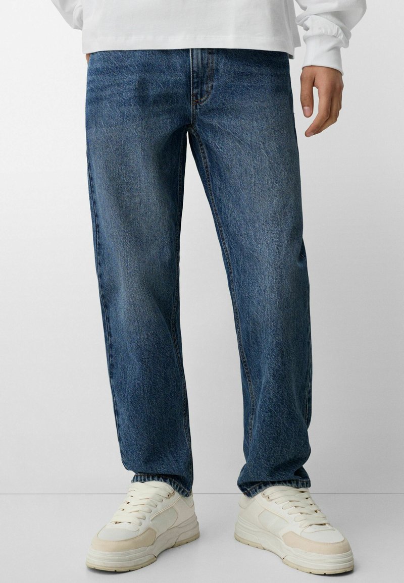Jeans in denim blu con una vestibilità rilassata, caratterizzati da zone sbiadite, design classico a cinque tasche e chiusura con zip. Abbinati a sneakers chiare.