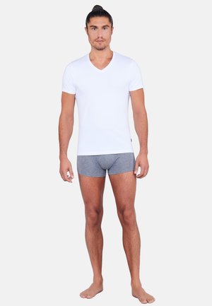 Mand med bundet hår, iført en hvid V-hals T-shirt og grå boxershorts, stående barfodet mod en ensfarvet baggrund.