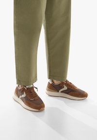 Sneakers marroni realizzati in materiali scamosciati e sintetici, con un elemento di design beige a contrasto. Le scarpe sono abbinate a pantaloni corti in olivastro.