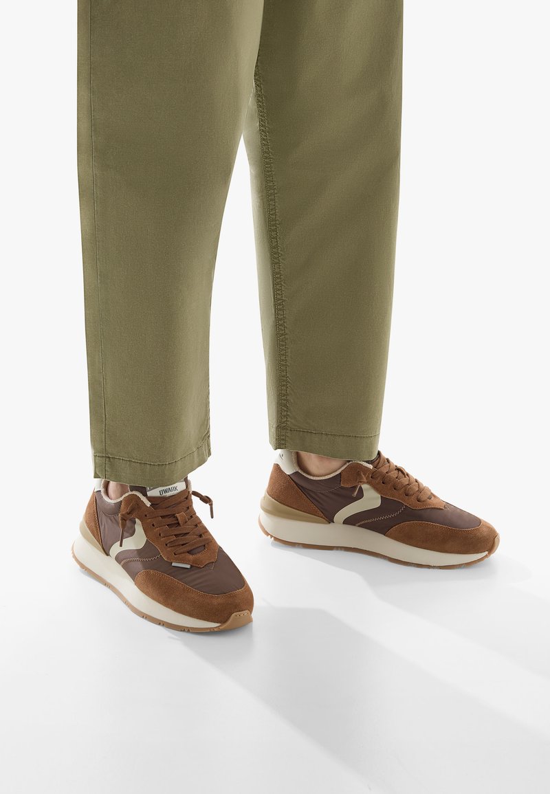 Sneakers marroni realizzati in materiali scamosciati e sintetici, con un elemento di design beige a contrasto. Le scarpe sono abbinate a pantaloni corti in olivastro.