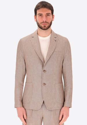 Uomo che indossa una giacca da completo in lino beige e pantaloni abbinati sopra una maglietta bianca con scollo rotondo, rivolto in avanti con espressione neutra.