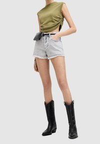 Top sin mangas de color verde oliva con fruncido lateral, pantalones cortos de mezclilla grises con dobladillo desgastado y botas de vaquero de cuero negro con diseño en relieve.