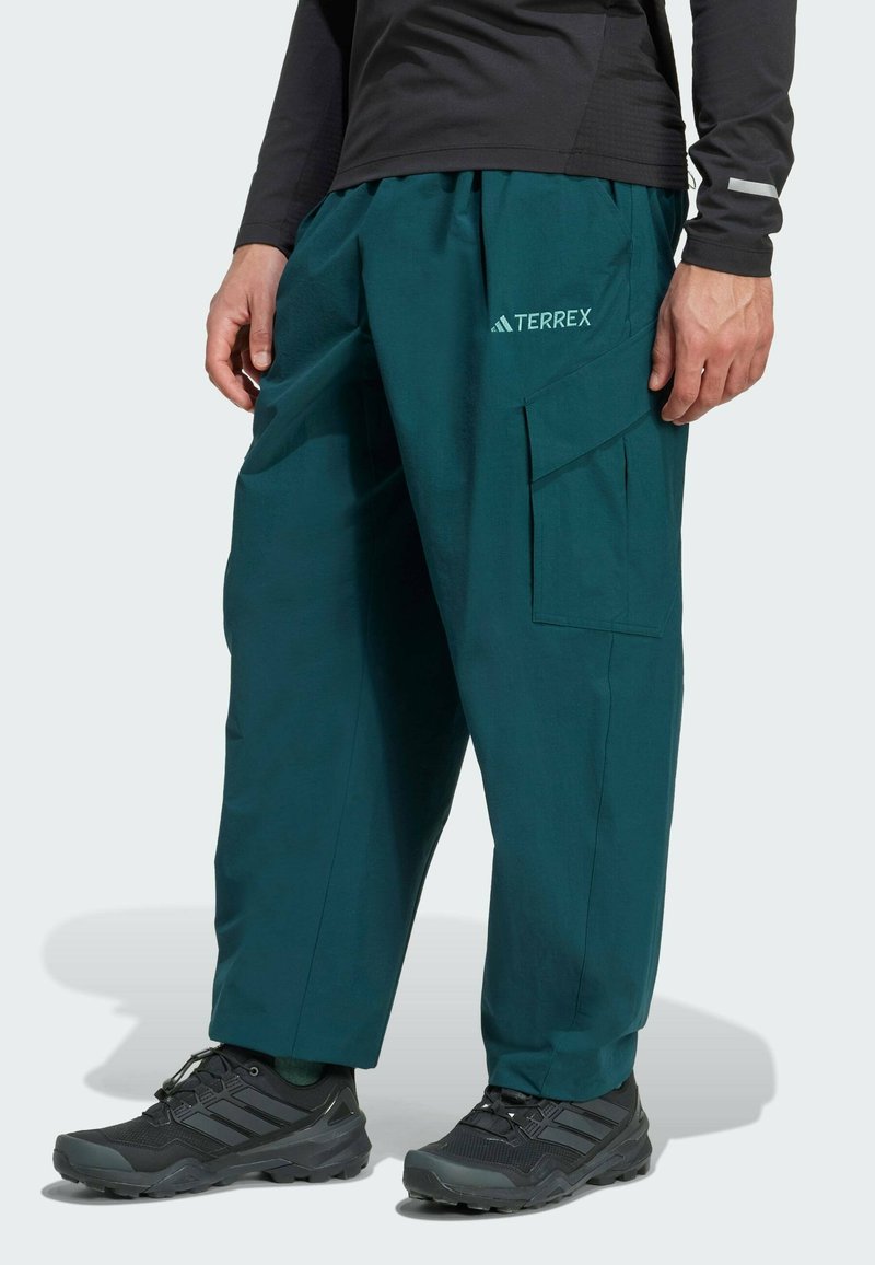 Teal udendørsbukser med cargo-lommer, elastisk talje og løs pasform. Sammen med sorte vandresko med teksturerede såler.
