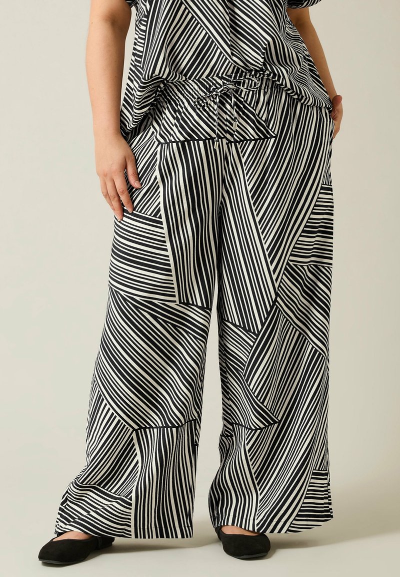 Personne portant un pantalon large rayé noir et blanc avec des motifs géométriques et des chaussures plates noires, debout les mains dans les poches.