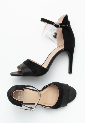 Een paar zwarte suède hoge hakken sandaaltjes met enkelbandjes en open tenen, één schoen met een zilveren 'Bull Boxer' merk binnenin.