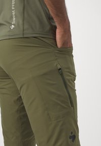 Olivgrüne Outdoor-Hosen mit einer seitlichen Reißverschlusstasche, glatter Textur und Logodetail. Verfügt über verstärkte Nähte und eine taillierte Passform.