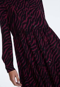 Langärmliges Kleid im dunkelroten und schwarzen Tigerstreifenmuster, mit Knopfleiste und gestuften Rüschen am Saum. Weiche Stoffstruktur.