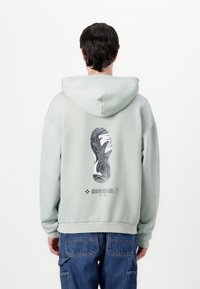 Hellgrüner Kapuzenpullover mit einem grafischen Druck einer Schuhsohle auf der Rückseite. Rippstrickbündchen und -saum, weicher Baumwollmischstoff.