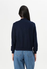 A woman with long hair wears a navy blue sweatshirt and light blue jeans, standing against a plain white background.