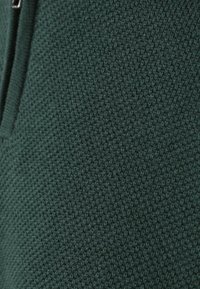 Donkergroene gebreide stof met een gestructureerd patroon, voorzien van een ritssluiting aan één kant. Het materiaal lijkt zacht en gestructureerd.