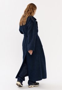 Una donna con lunghi capelli mossi indossa un lungo cappotto blu scuro, jeans scuri e sneakers, mentre cammina su uno sfondo bianco semplice.
