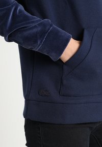 Hand i fickan på marinblå sweatshirt med texturerad ärm och liten broderad krokodil-logotyp nära nederkanten.