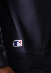 Primer plano de la manga y el dobladillo de una sudadera oscura que presentan un pequeño parche con el logo de la MLB, con la silueta de un bateador en azul, blanco y rojo.