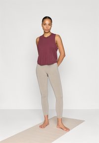 Mørkerød ærmeløs top parret med beige højtaljede leggings. Blødt stof og tætsiddende design. Stående på et lys beige yogamåtte.