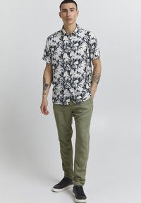 Chemise blanche à manches courtes avec des motifs floraux et des feuilles noires, portée avec un pantalon vert olive et des baskets noires avec des accents argentés.