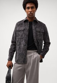 Chemise à boutons noire à motifs floraux, avec un col, deux poches poitrines et des manches longues. Portée par-dessus un pull noir et un pantalon gris.