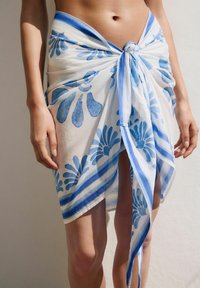 A model wears a light, floral-patterned wrap skirt with blue accents, showcasing a relaxed summer style against a neutral background.