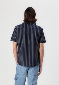 Homme aux cheveux mi-longs portant une chemise bleu marine à manches courtes avec des rayures verticales blanches et un jean bleu clair, debout de dos.
