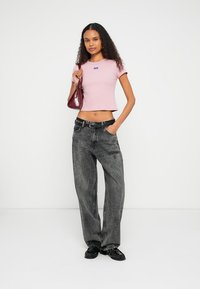 Rosa kort t-shirt med logga, kombinerad med lösa svarta denimjeans. Accessoarerna inkluderar en liten vinröd handväska och svart fotbeklädnad.