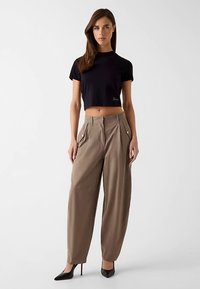 Schwarzes, kurz geschnittenes T-Shirt mit kurzen Ärmeln und Logo; beige, weite Hose mit Bundfalten und goldenen Knopfdetails; schwarze High Heels.