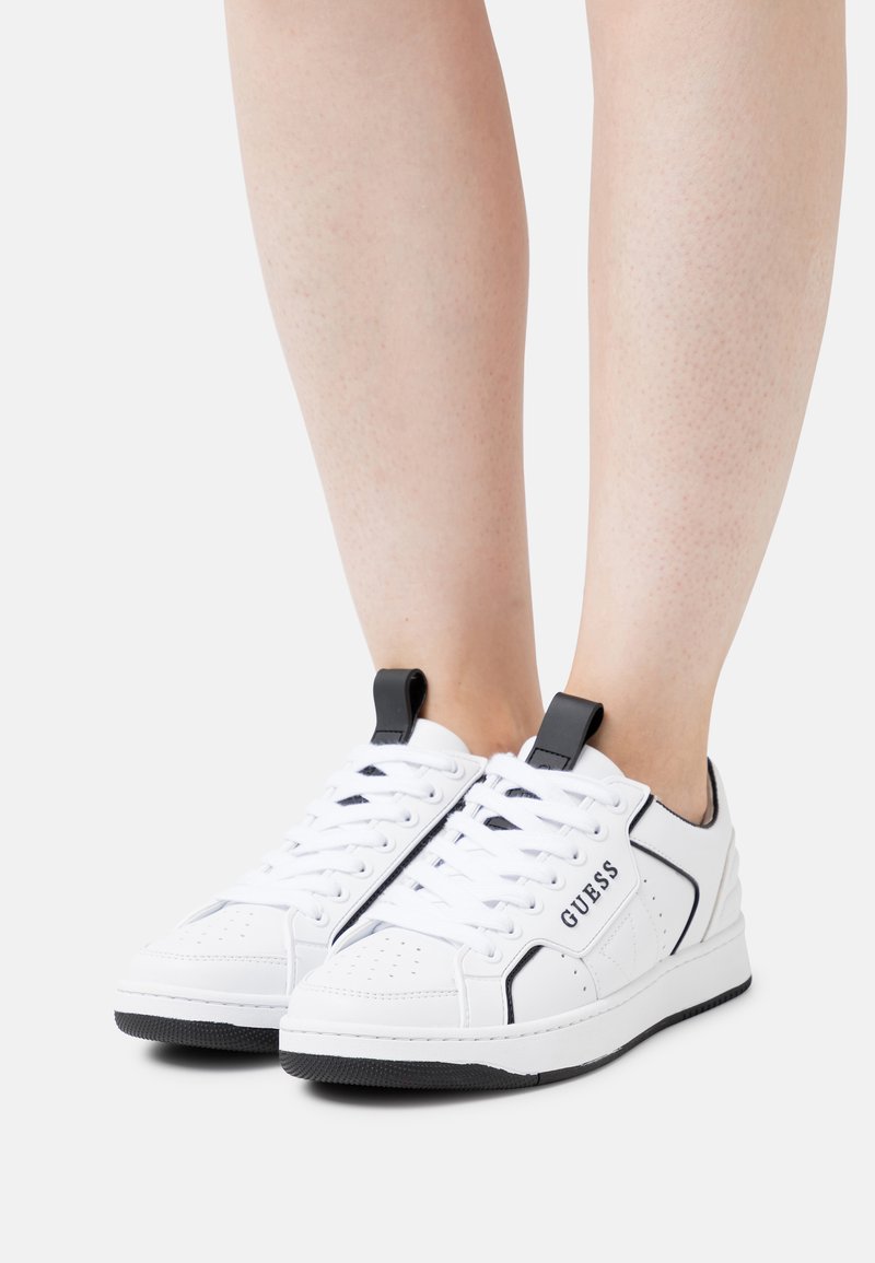 Sneakers bianchi con accenti neri, parte superiore in pelle e tessuto, design con lacci, puntale perforato e suola in gomma nera. Logo "GUESS" presente.