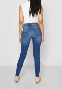 Jeans ajustados de mezclilla en color azul, con una cintura alta, dos bolsillos traseros y un parche de cuero marrón en la cinturilla. Textura suave.