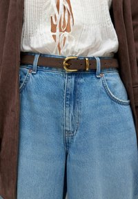 Vitt texturerat blus med snörning framtill, brun mockabälte med guldfärgad spänne, blå högmidjade jeans, och bruna ärmar av jacka synliga.
