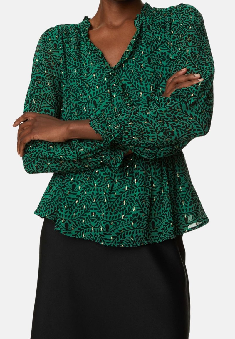 Blouse verte à motifs avec un bas évasé, des manches longues et un décolleté en V. Le tissu semble léger, avec des accents dorés dans le design.