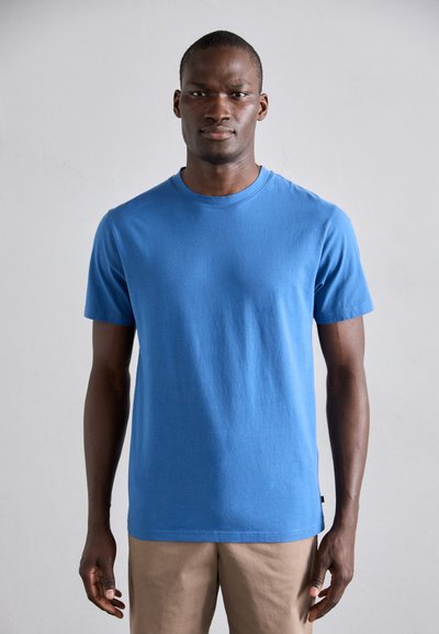 Blaues Baumwoll-T-Shirt mit kurzen Ärmeln, Rundhals und glatter Textur. Wird mit hellbeigen Hosen getragen, minimale Markenkennzeichnung am Saum sichtbar.