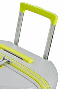 Valise rigide gris clair avec des accents jaunes vifs. Comprend une poignée rétractable et des doubles zips avec une prise texturée.