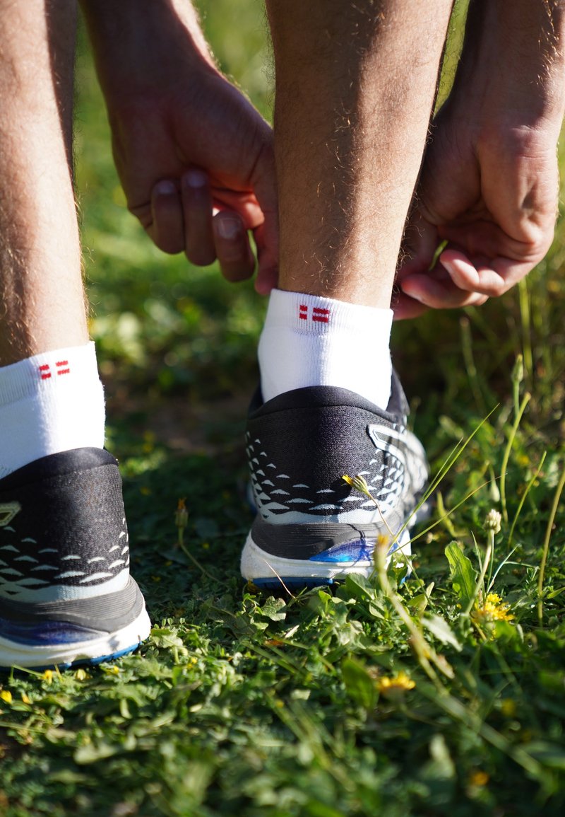 Zapatillas deportivas negras con acentos blancos; calcetines cortos blancos con diseños rojos; manos ajustando las mangas; fondo de césped con flores.