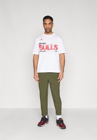 Λευκό υπερμέγεθες t-shirt με κόκκινο γράφημα "BULLS", πράσινο παντελόνι με στενή γραμμή και μαύρα/κόκκινα αθλητικά παπούτσια. Το μοντέλο στέκεται μπροστά σε απλό φόντο.