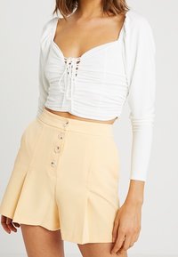 White cropped top with lacing detail and long sleeves paired with high-waisted pale yellow shorts featuring three front buttons and pleats.
