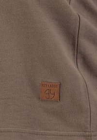 Brauner Stoff mit einer glatten Textur präsentiert ein quadratisches Lederpatch mit der Aufschrift "KEY LARGO" und einem Logo. Das Design enthält klare, einfache Linien.